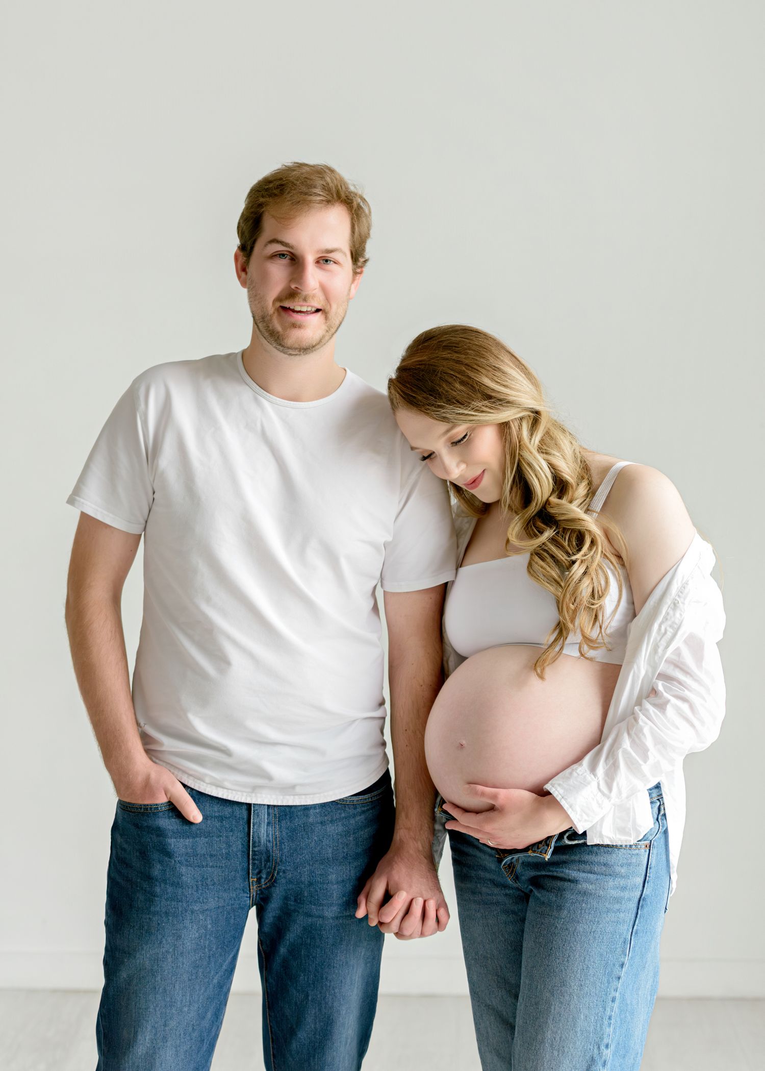 Expecting couple in white shirts and jeans share tender poses against light background in maternity photo series.
