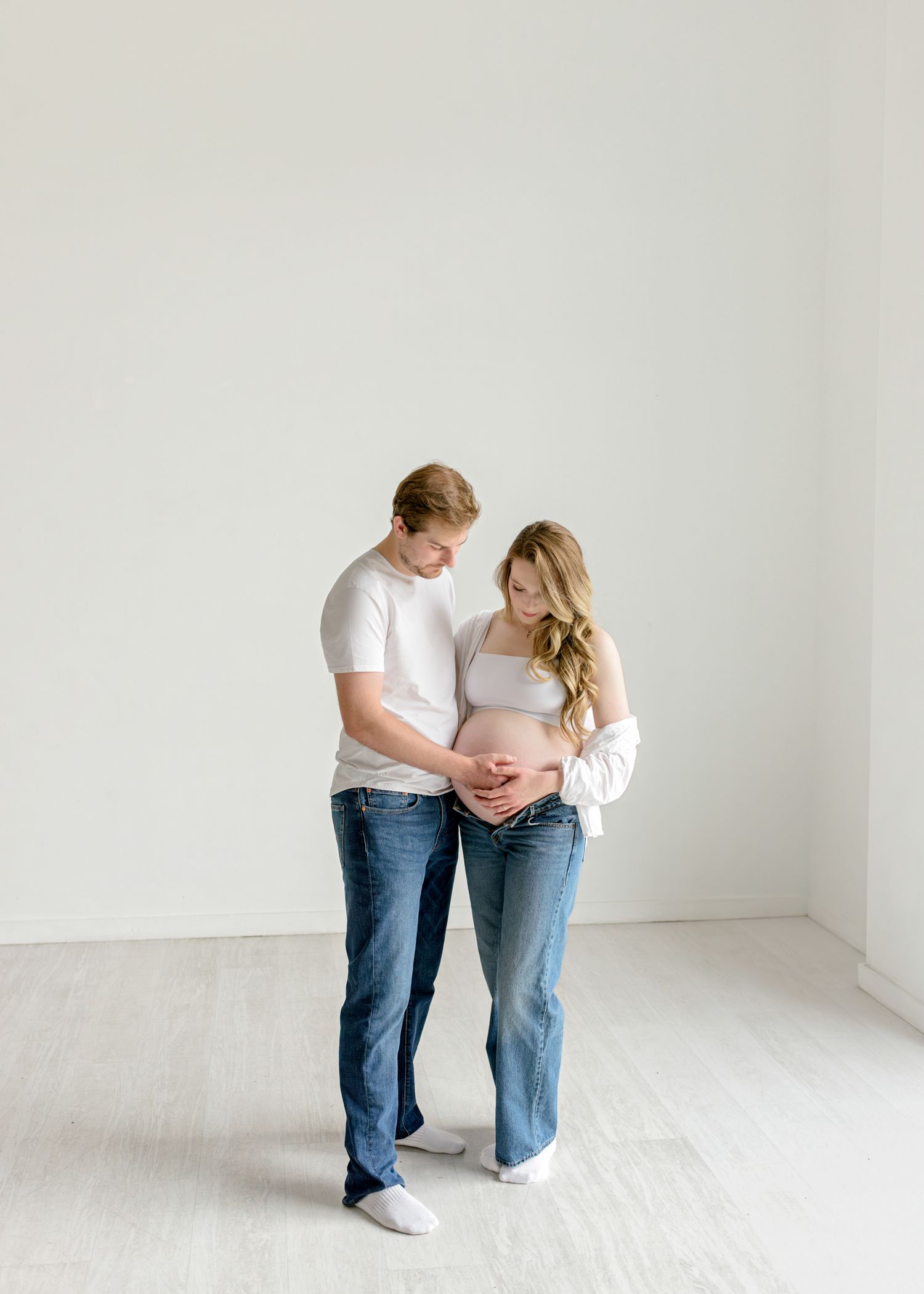 Couple in white shirts and jeans embrace while gazing at her pregnant belly against a minimal white backdrop.