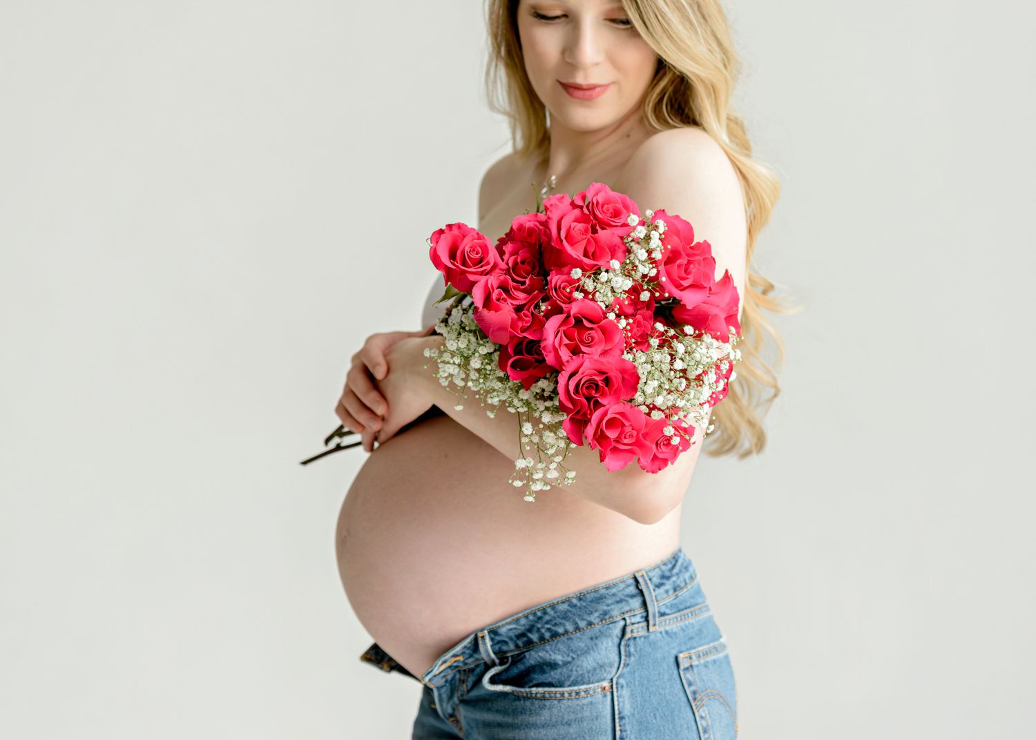 Side view of an expectant mother holding a bouquet of vibrant pink carnations against a white backdrop.
