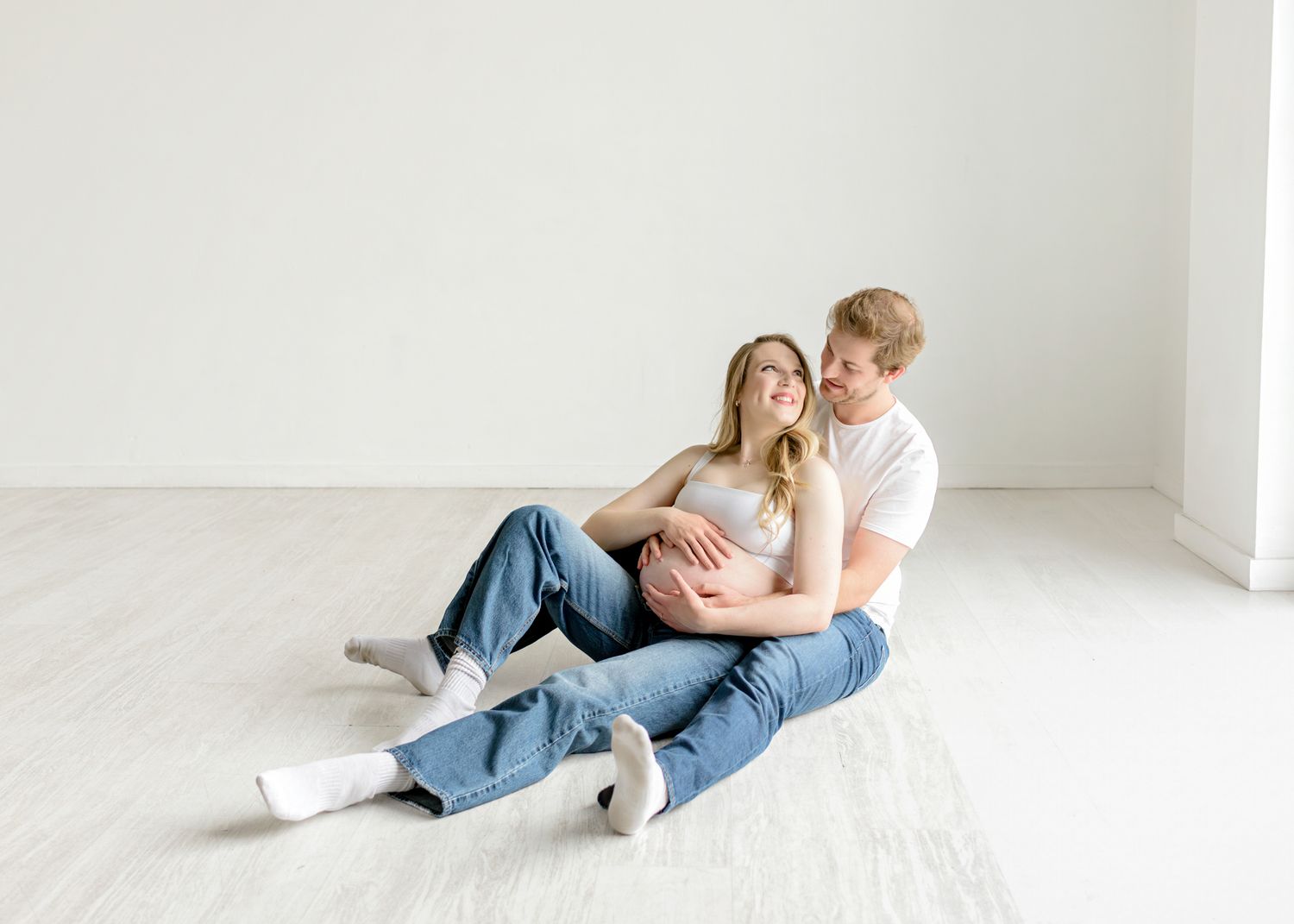 A couple in matching denim sits together on a white floor in a bright, minimalist room.