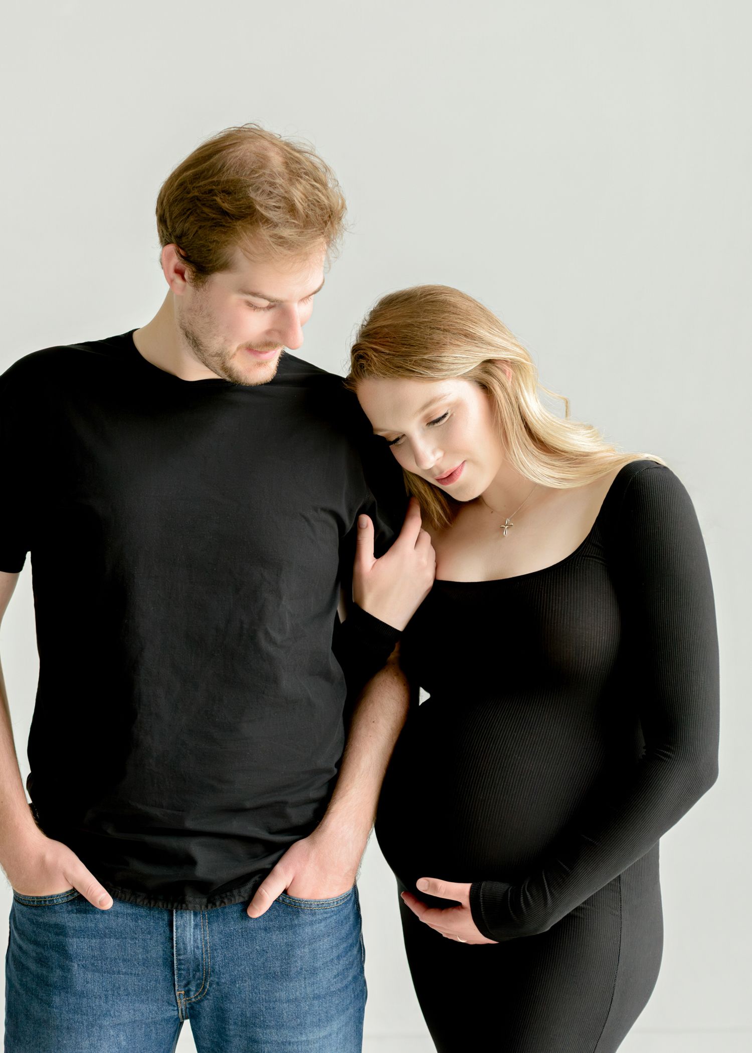 Intimate maternity photo of expecting couple in black outfits against light background.