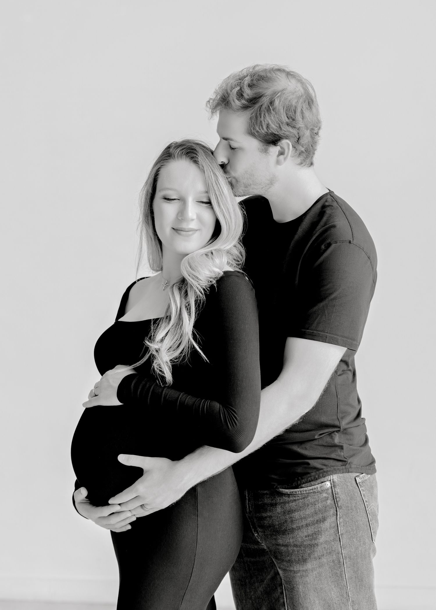 Black and white maternity portrait of couple embracing with man kissing woman's forehead.