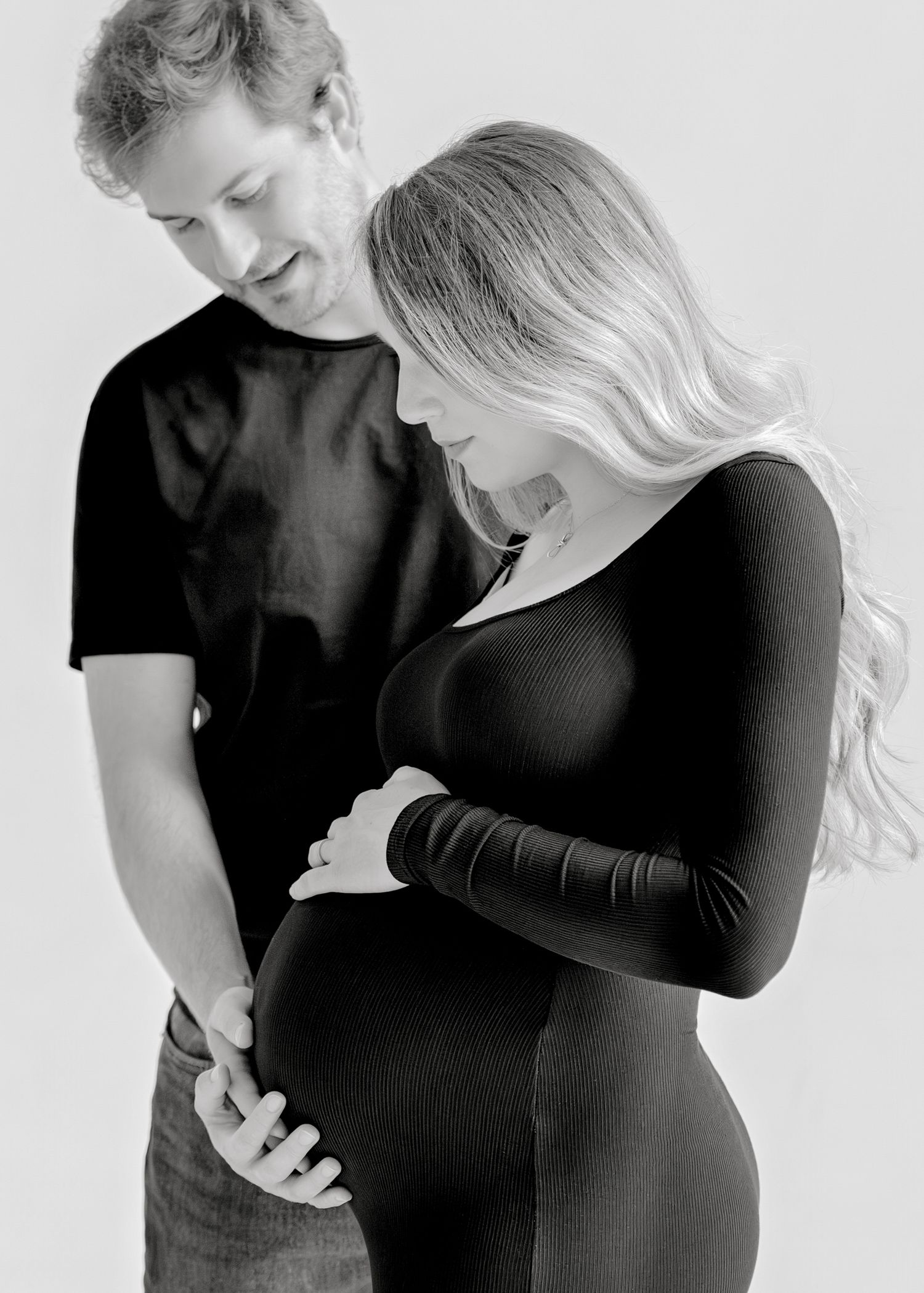 Black and white maternity photo showing a couple in dark clothing embracing and focusing on her pregnant belly.