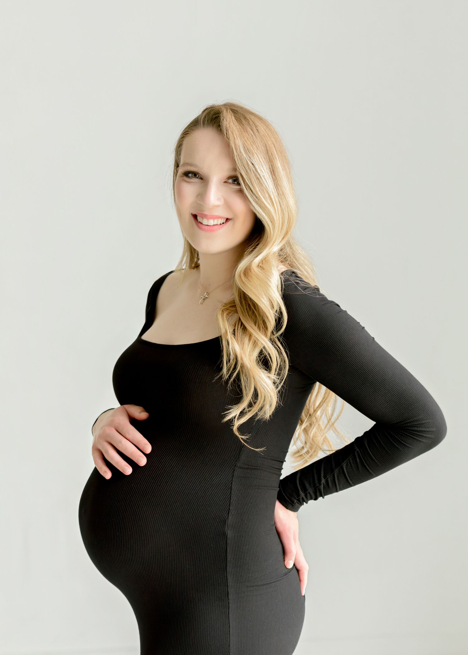 Woman in black long sleeve dress smiles while cradling her pregnant belly against light background.