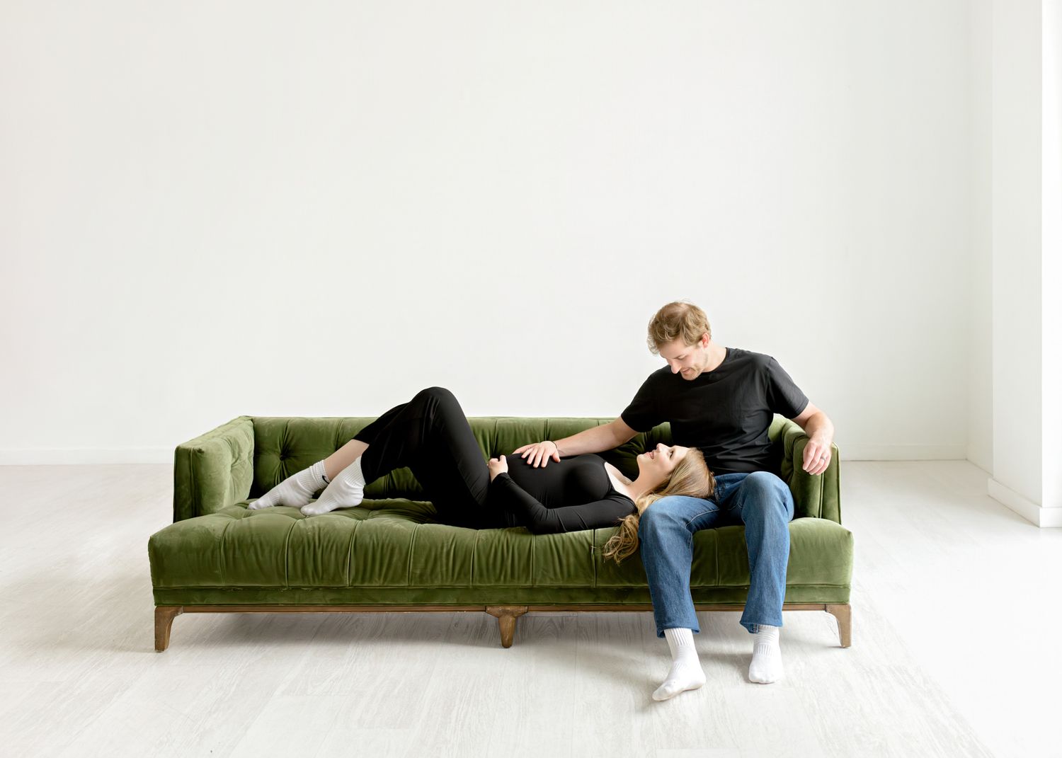 Someone relaxes on a plush green velvet sofa while reading in a minimalist white room.