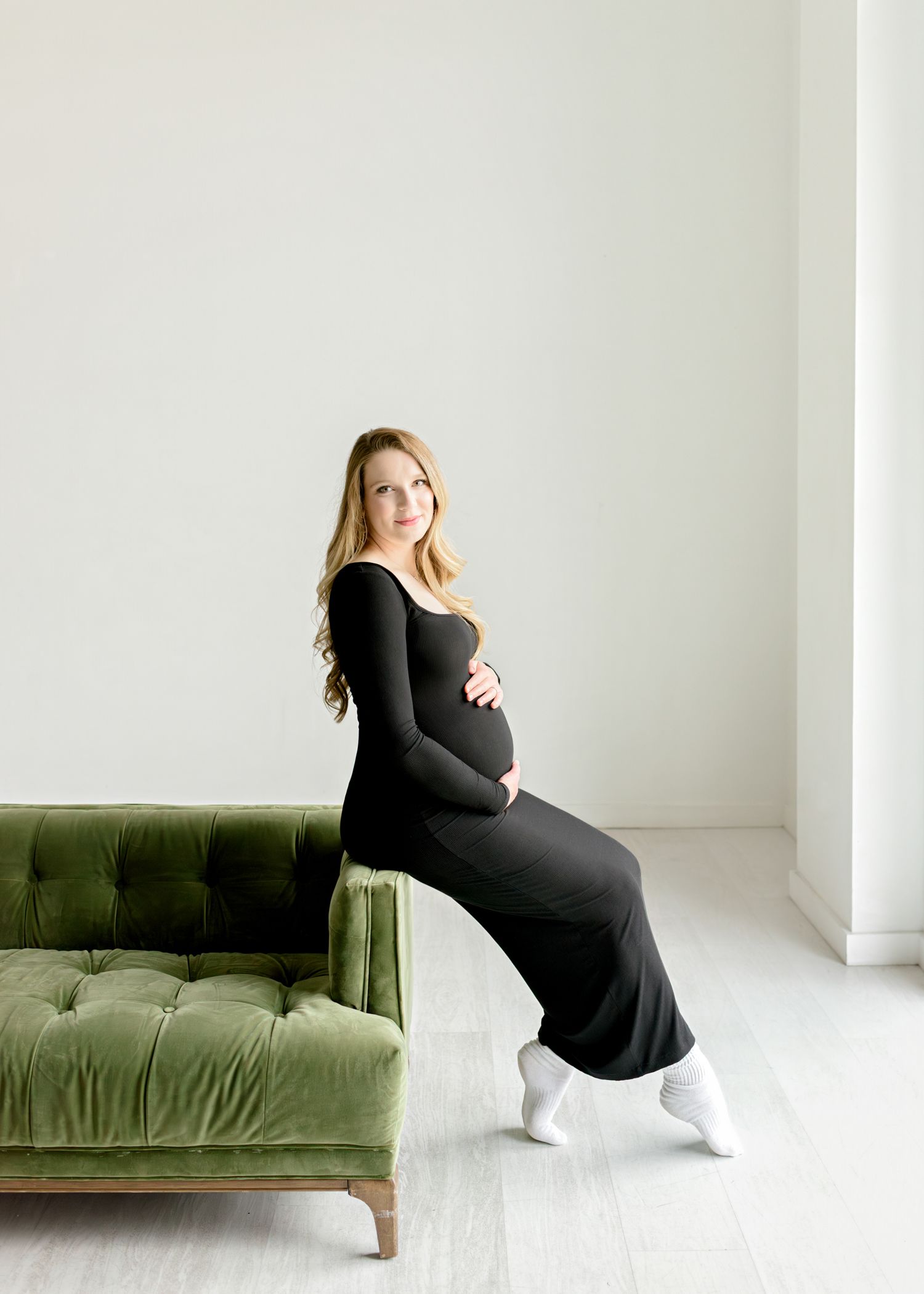 Expecting mother in black dress sits on olive green velvet sofa in minimalist white room while wearing white sneakers.