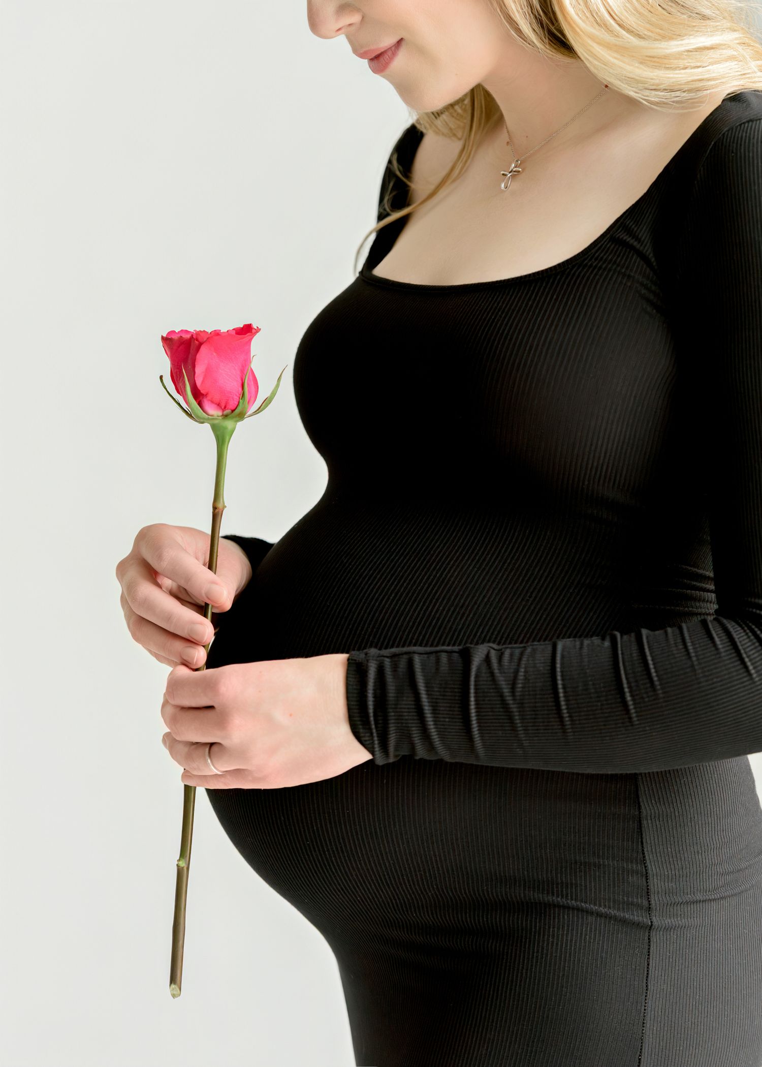 Side view of pregnant woman in black dress holding single red rose against white background.