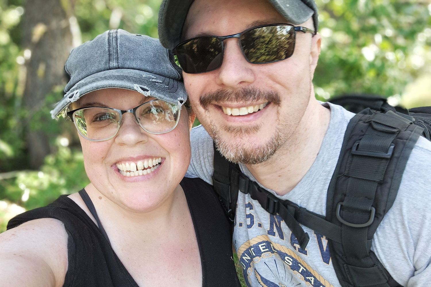 A couple wearing casual outdoor attire with hats and sunglasses take a selfie together on a sunny day.