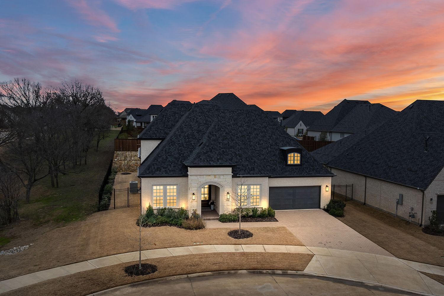 Modern suburban home with gray shingle roof and warm interior lighting at dusk with colorful sunset sky.