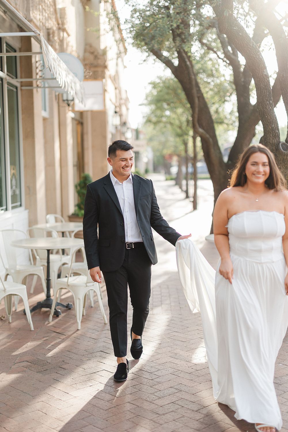 A couple holding hands and walking together down a tree-lined sidewalk outside a Mediterranean-style building.