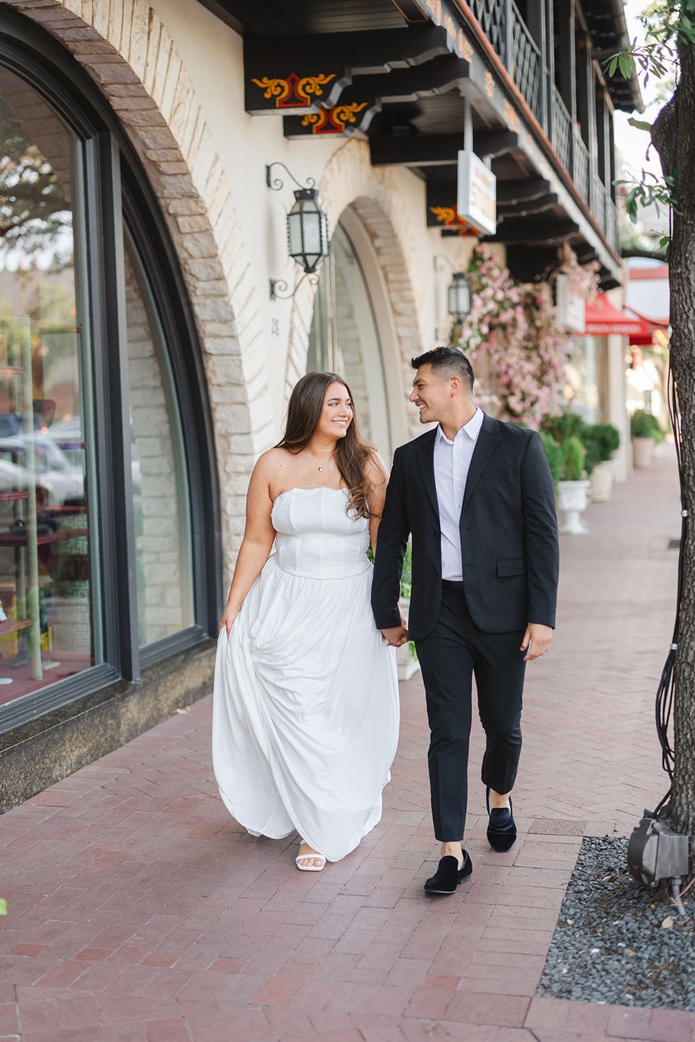 Couple in wedding attire walking along a Spanish-style building with arched walkways and terracotta tiles.