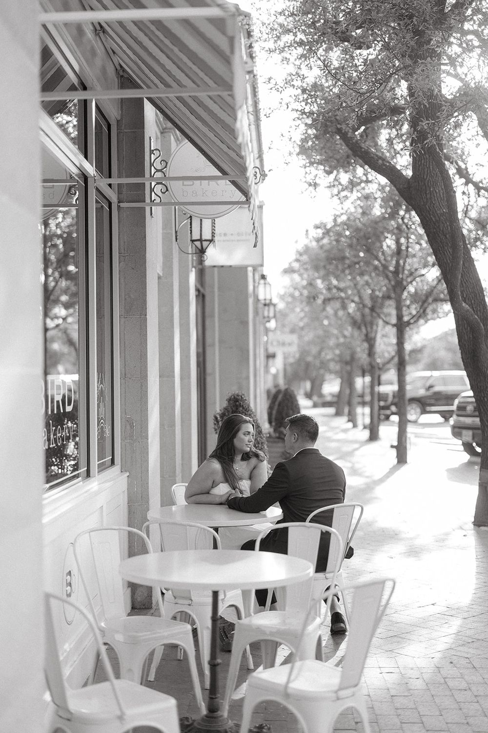 People enjoying coffee at outdoor cafe tables along a sidewalk with trees in black and white photography.