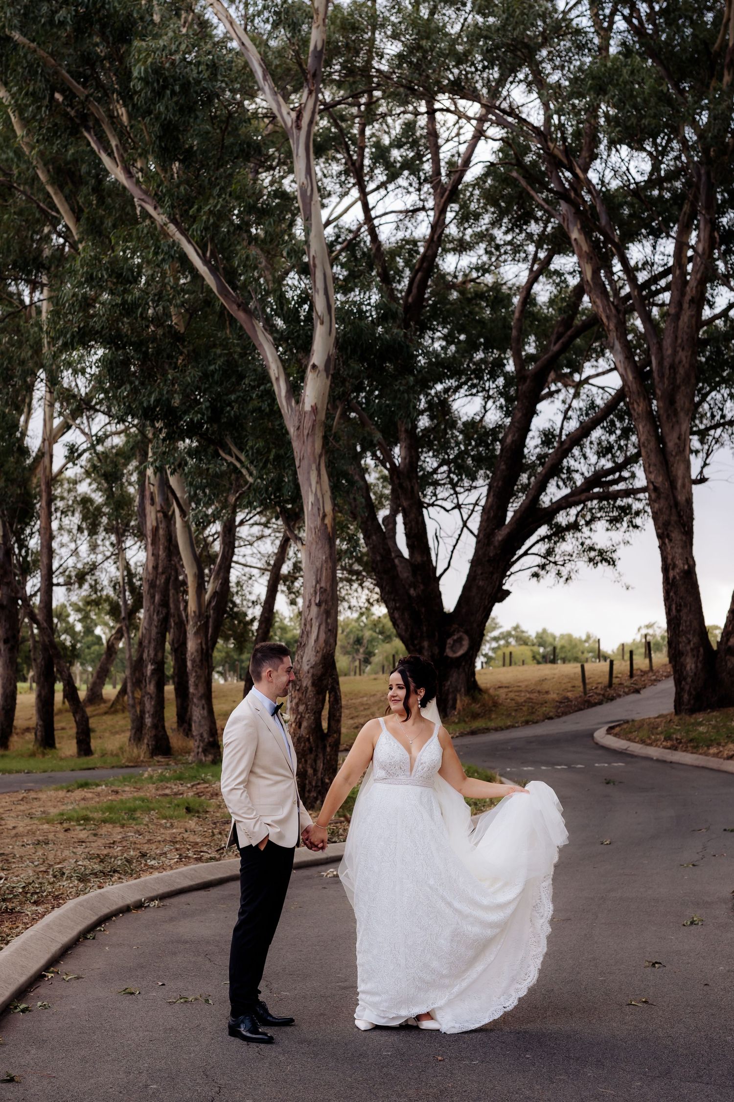 A couple stands on a winding path under tall eucalyptus trees, with the bride twirling her white gown.