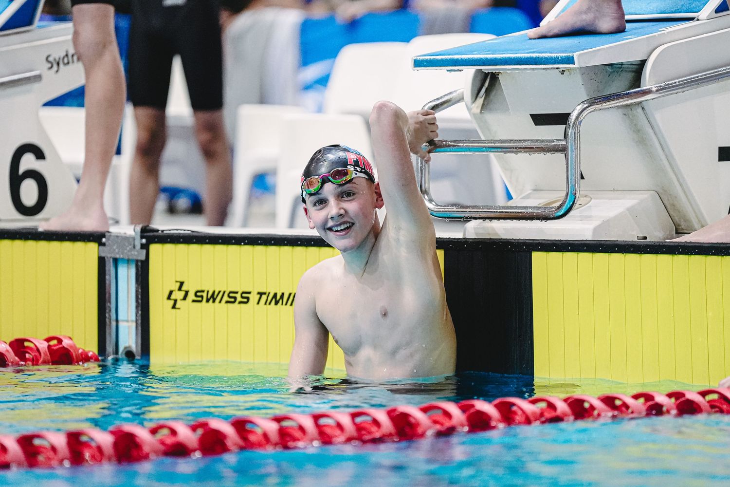 A swimmer competes in a competitive race at a swimming pool with timing devices visible in the background.