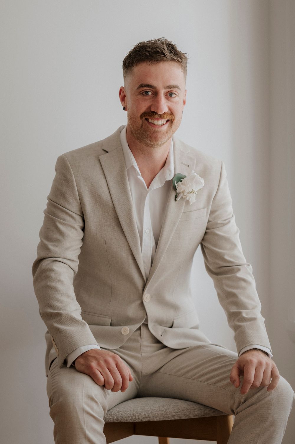 Groom poses in a light beige suit against minimal white background.