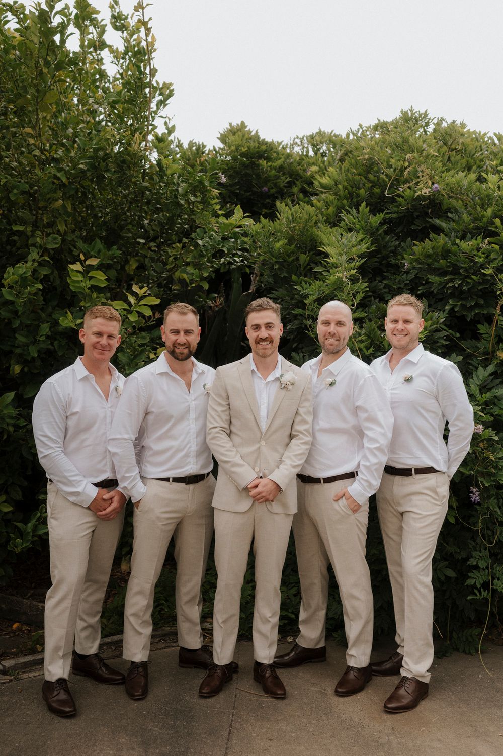 Groomsmen in coordinated beige and white attire pose together against garden backdrop.