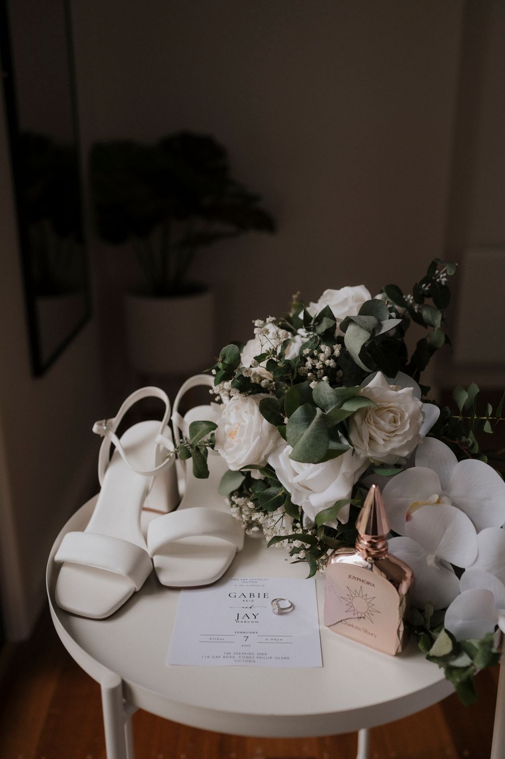 White bridal shoes and wedding stationery arranged with roses and greenery on a table.