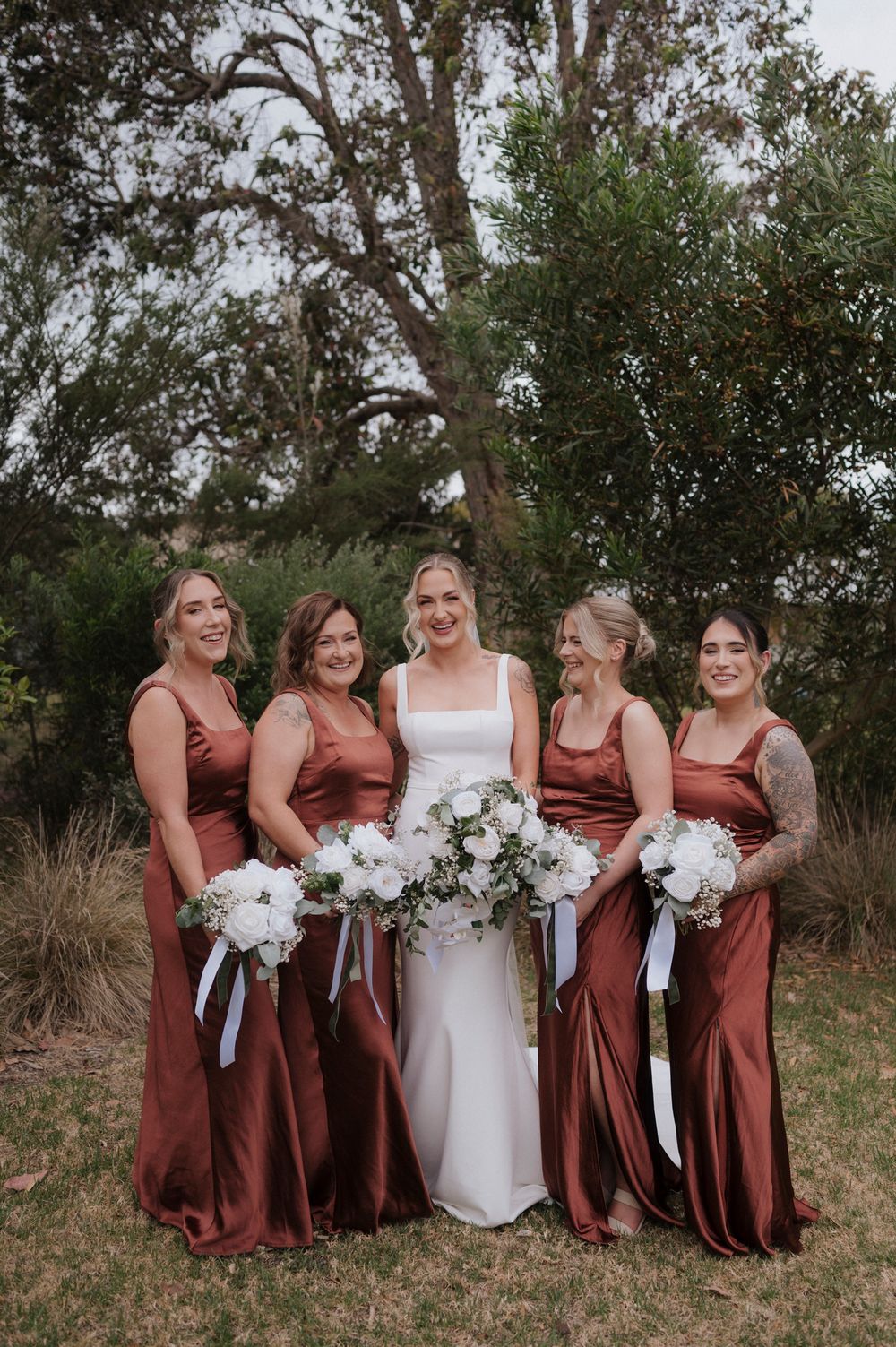 Bridal party in rust-colored dresses pose together outdoors.