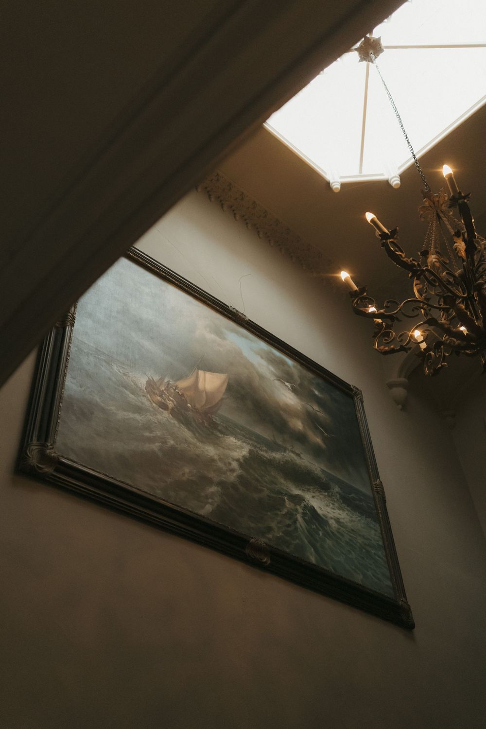 A dramatic seascape painting hangs on a wall beneath a chandelier in moody lighting.