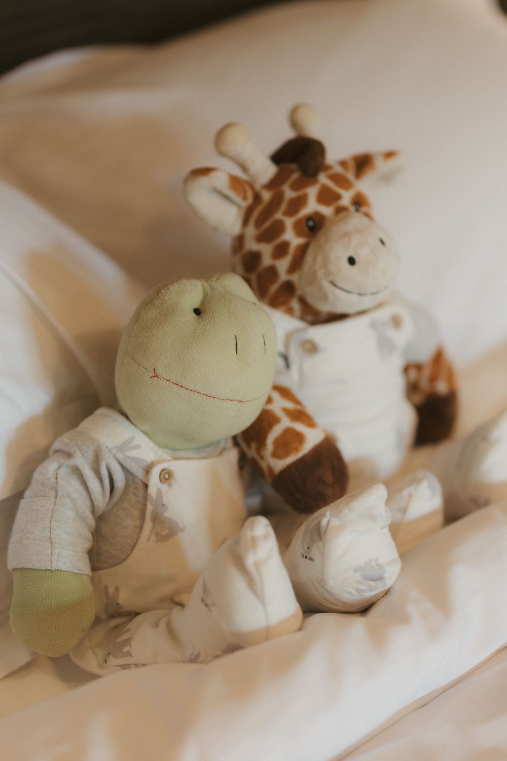 Soft plush toys of a giraffe and frog arranged on white bedding.
