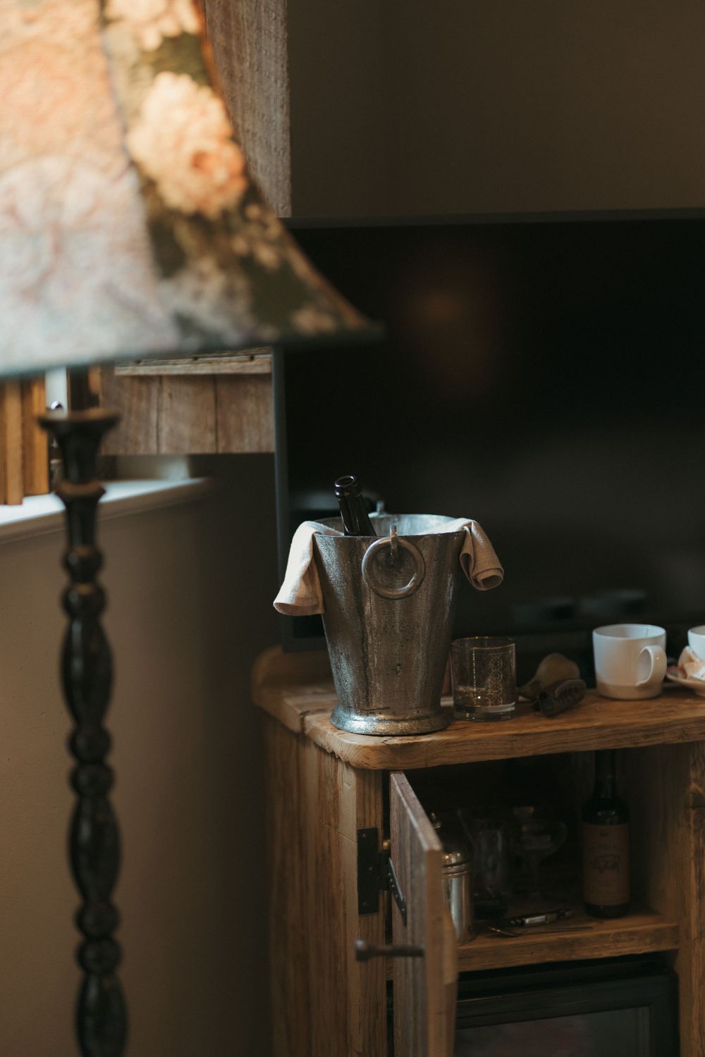 A vintage wooden side table with an ice bucket and glasses sits under a decorative lamp in a dimly lit room.