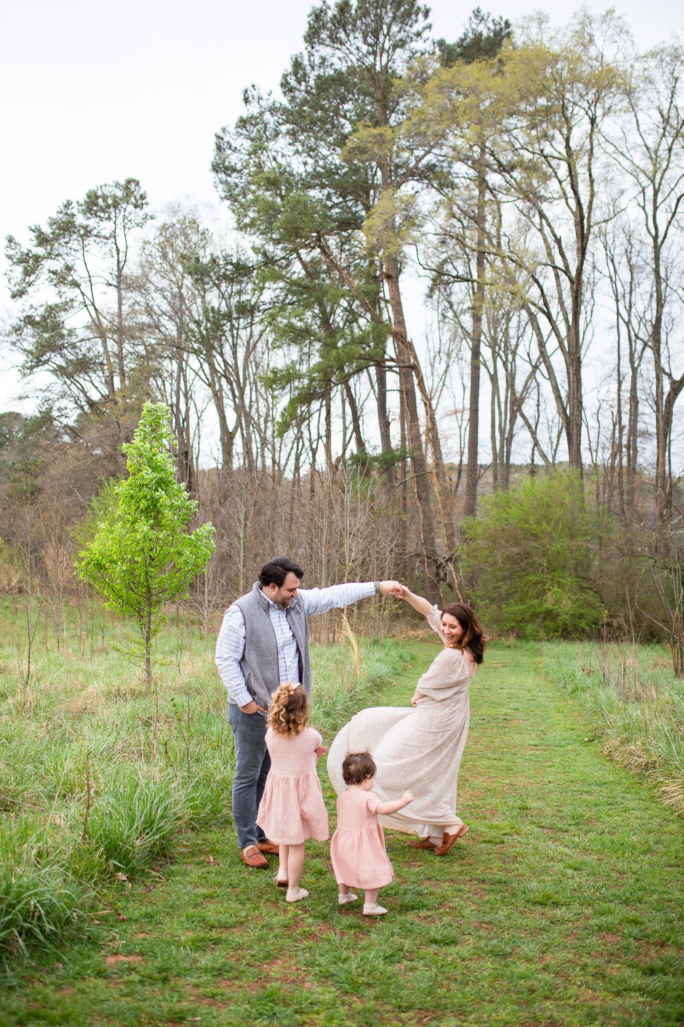 A family enjoys a playful dance moment together in a grassy field surrounded by tall trees on a spring day at Sugg Farm