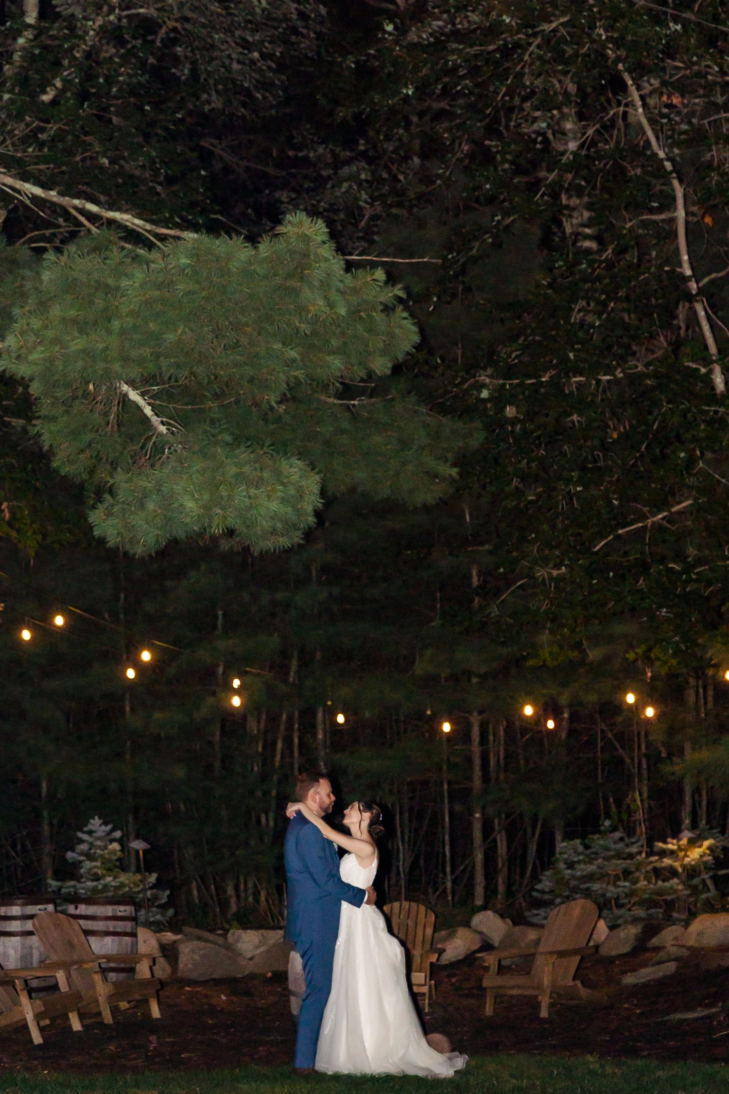 A romantic evening scene with string lights illuminating Adirondack chairs and tall trees in the background.
