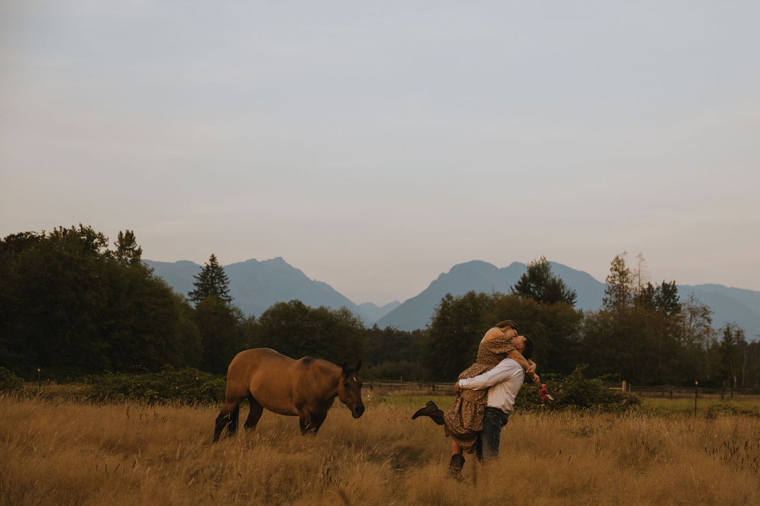 Washington Couple embracing in a golden grass field at sunset while a brown horse walks nearby and mountains in the background.