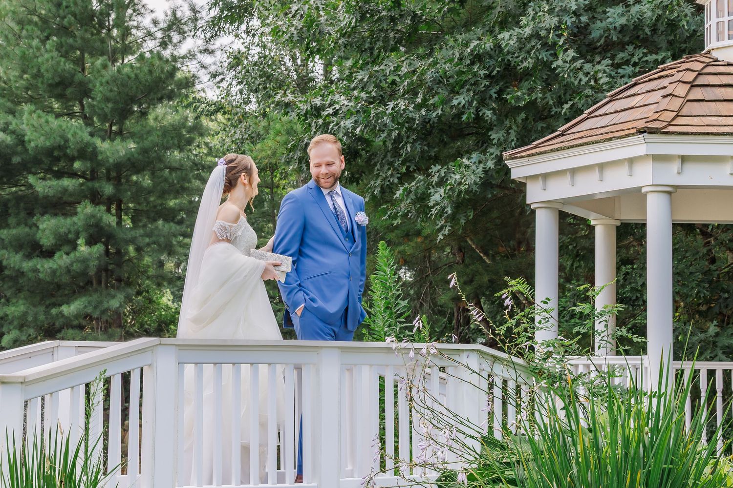 Happy couple embraces and smiles together on an elegant white gazebo deck surrounded by lush green trees.