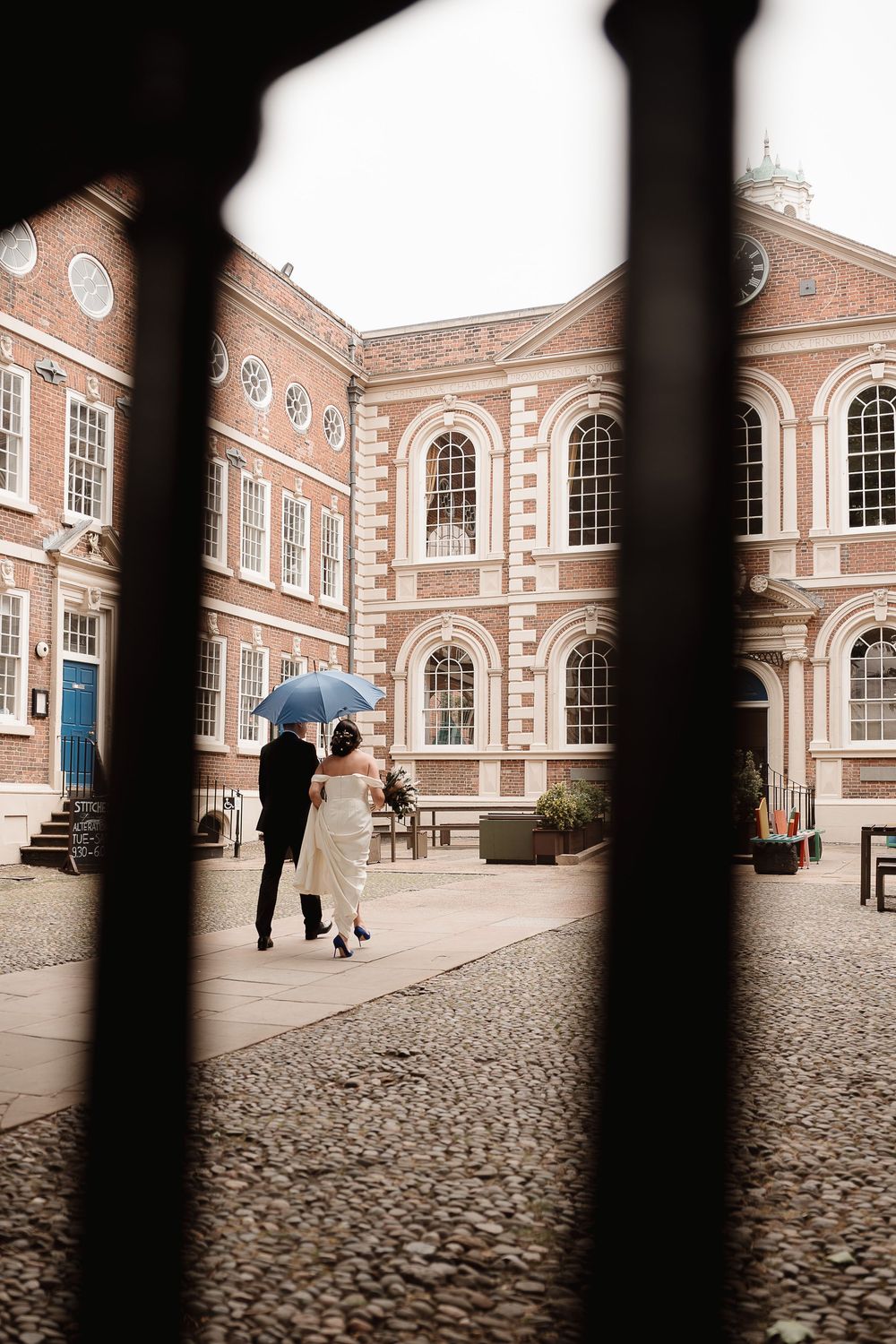 Two figures with umbrella walk across cobblestone courtyard of elegant Georgian brick building with arched windows.
