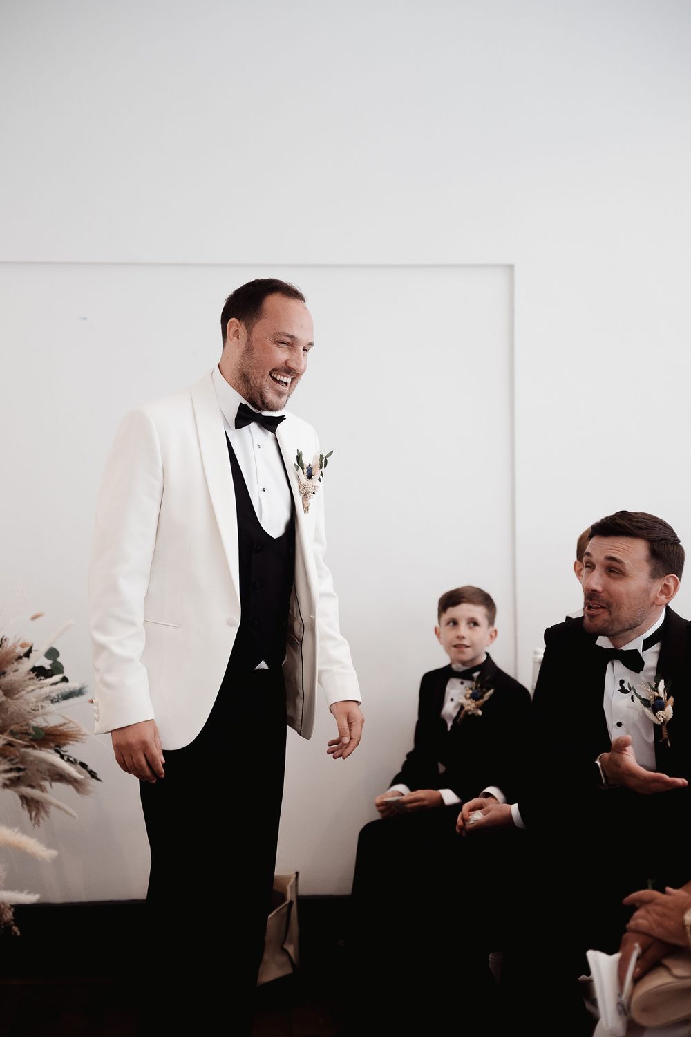 Wedding party members in white tuxedo jackets and black vests share candid laughter during formal photos.