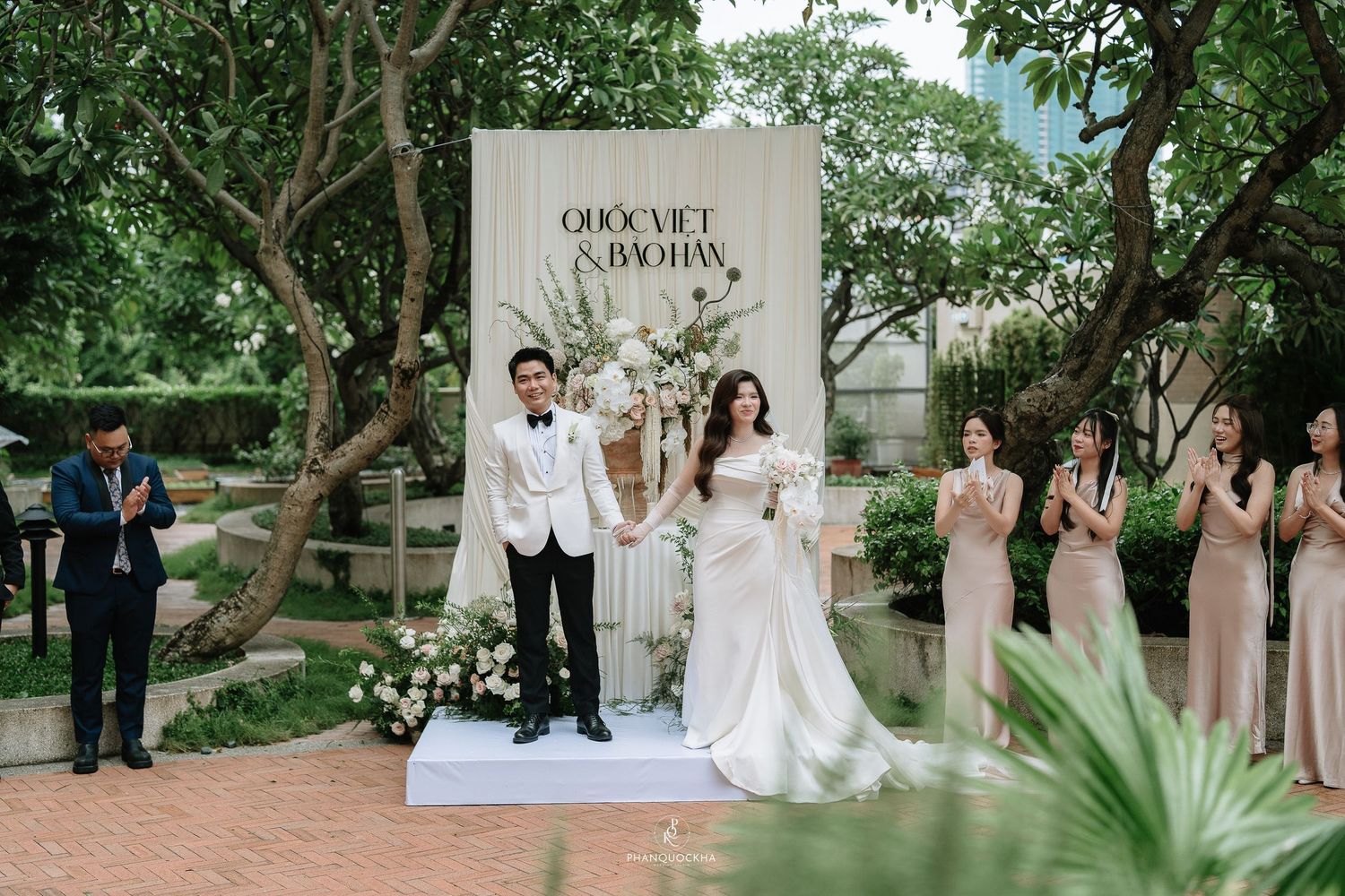 Bride and groom celebrate at an outdoor garden wedding ceremony with bridesmaids and guests applauding.