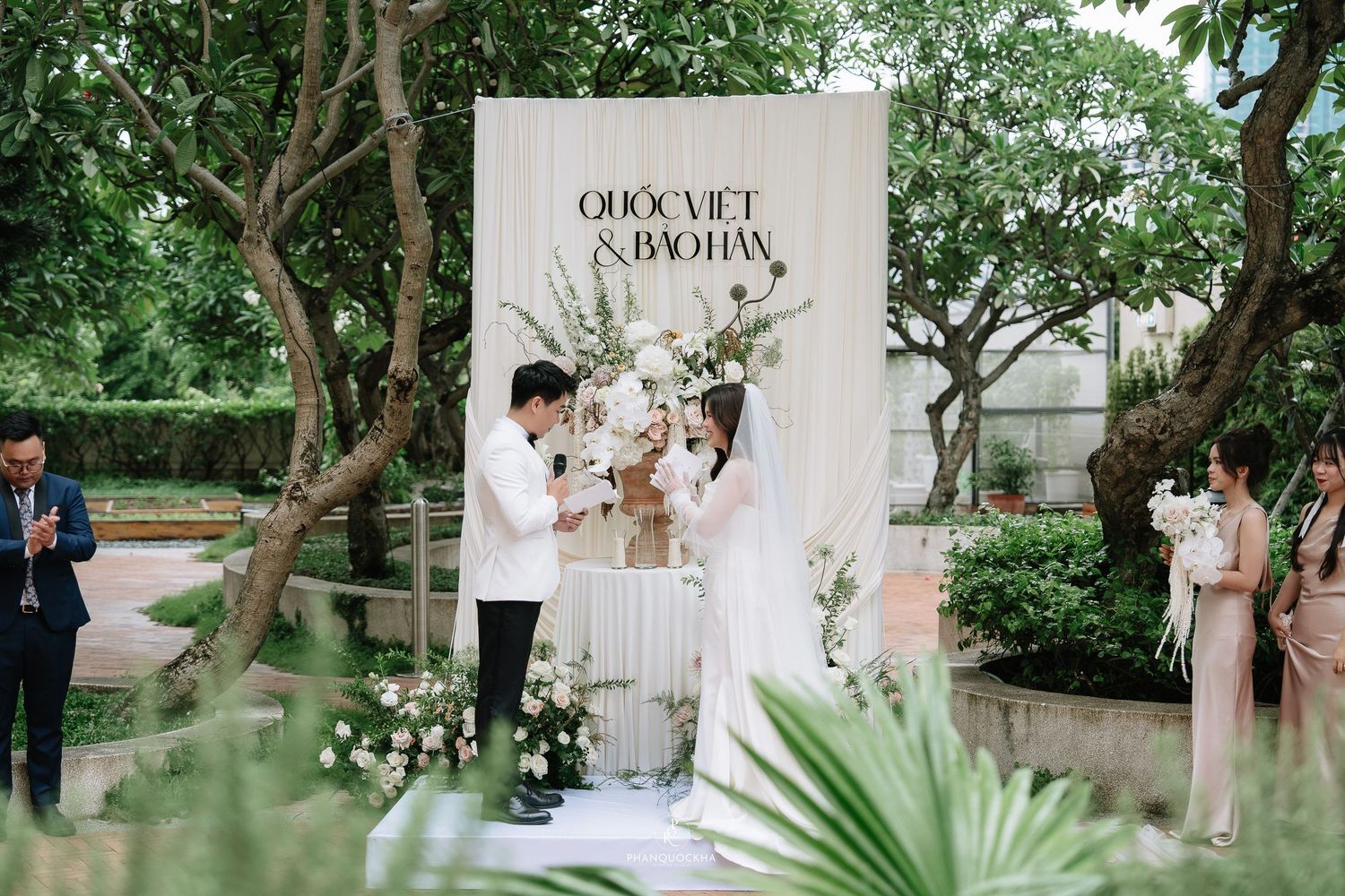 Bride and groom exchange vows at outdoor wedding altar decorated with white flowers and draped fabric backdrop.