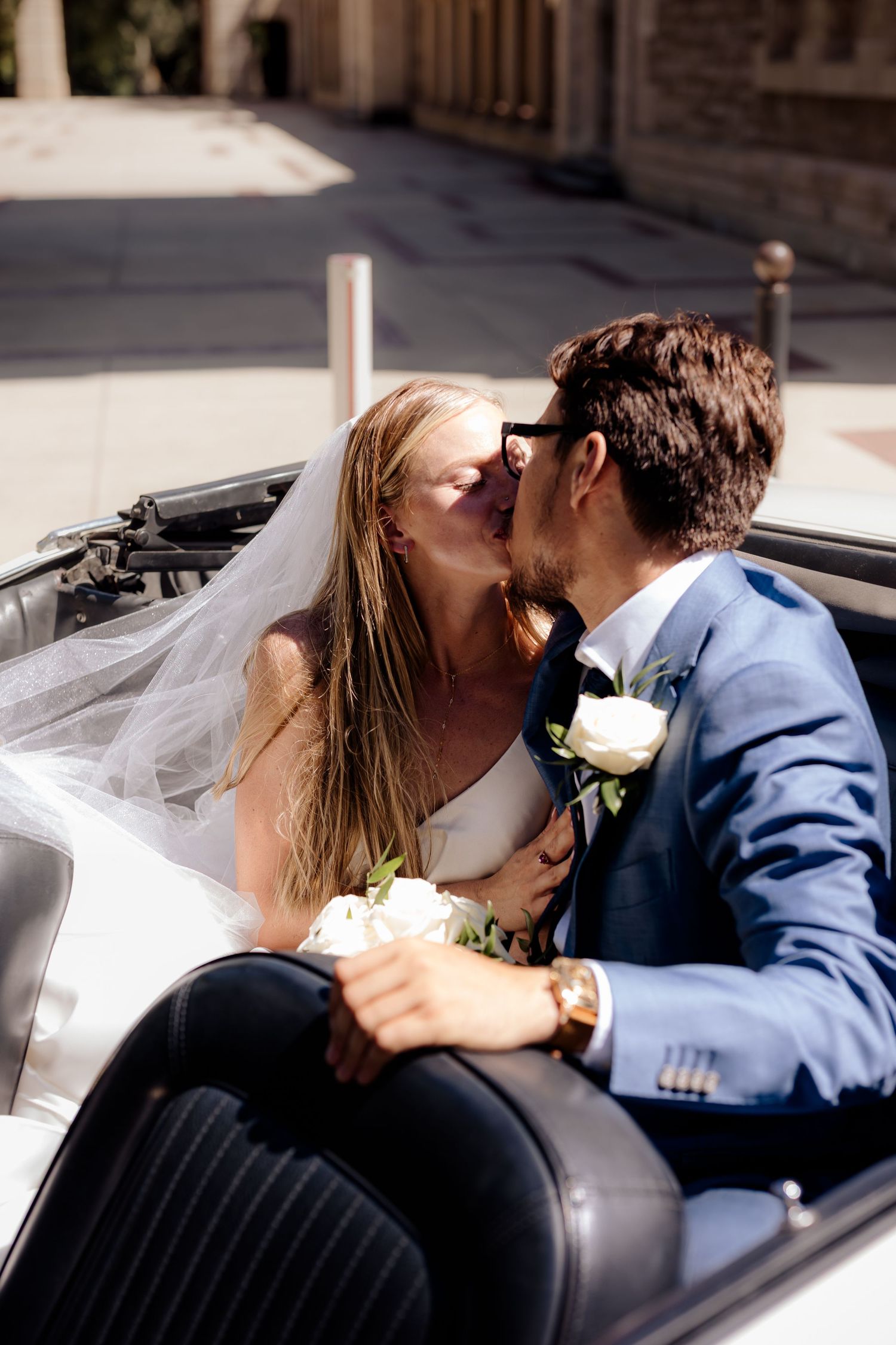 A newly married couple shares romantic kisses in the front seat of a convertible car during their wedding photoshoot.