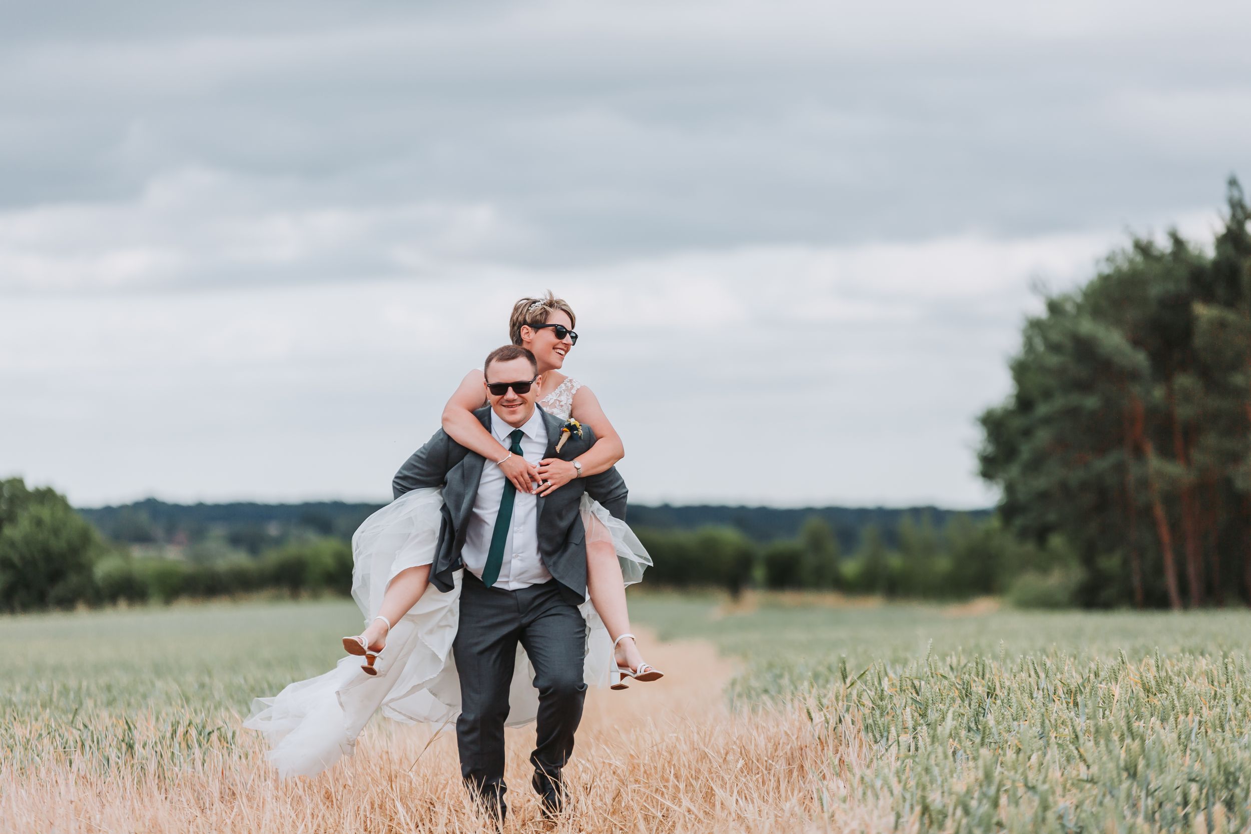 Newlyweds dance and celebrate together in a sunlit meadow with tall grass and evergreen trees in background.