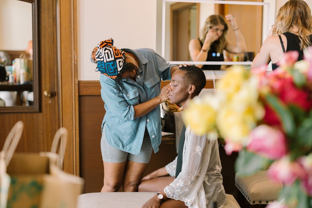Candid moment of bride getting makeup done by friends during wedding prep at Geneva Terrace, with colorful flowers in the foreground.