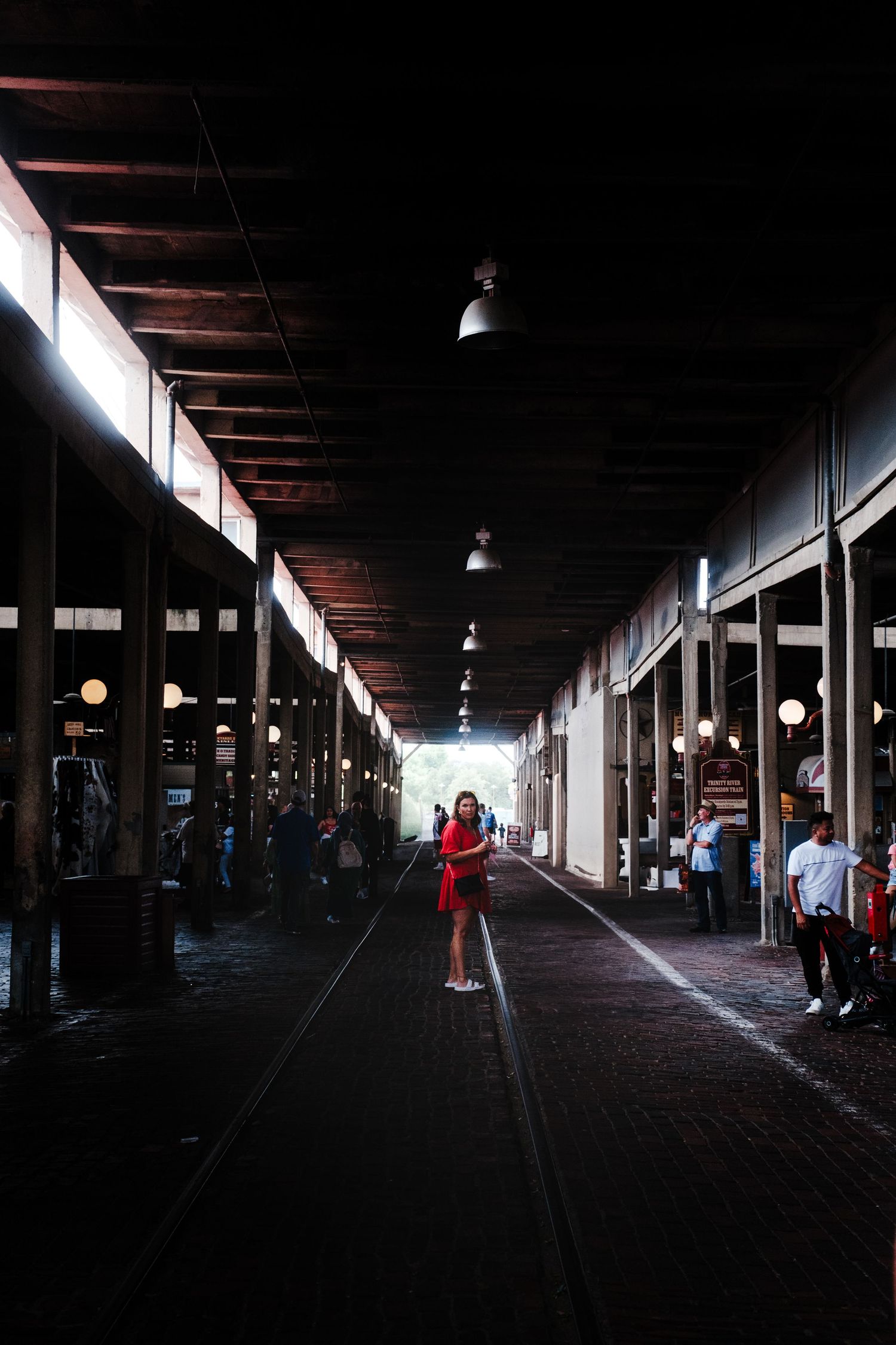 Figure in red dress stands in a dark covered arcade walkway with dramatic lighting and columns on either side.