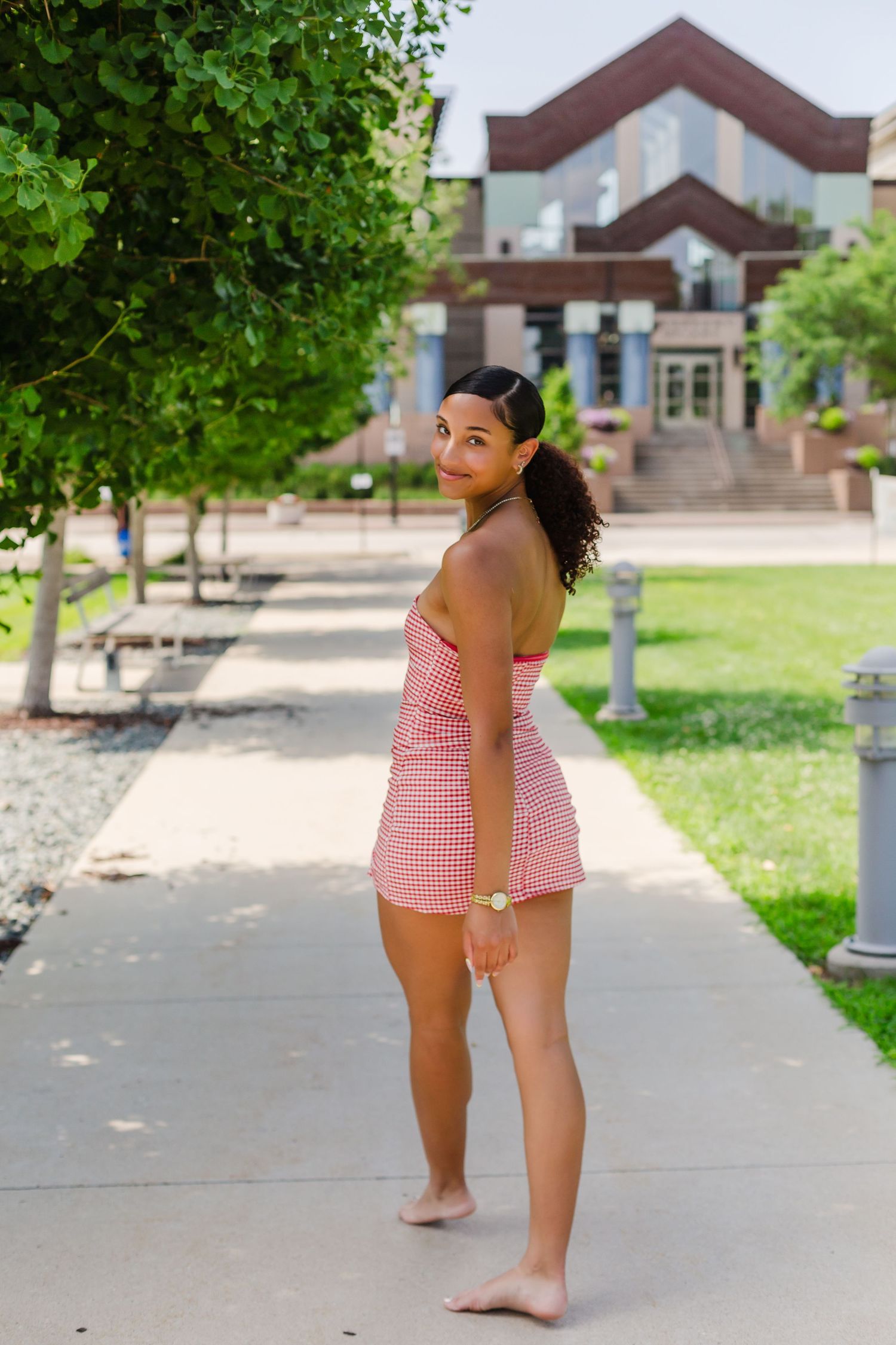 A child in a pink gingham swimsuit plays on a sunny sidewalk with trees and buildings in the background.