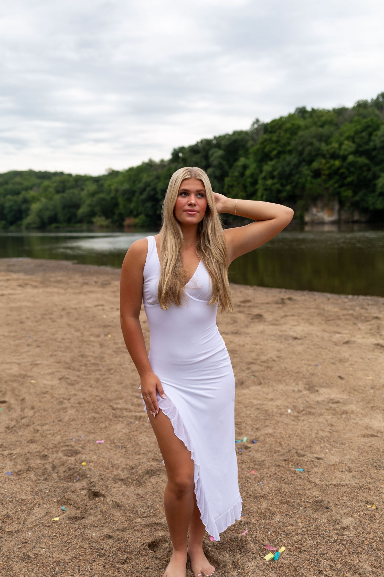 Woman in white dress poses on sandy beach near lake and wooded hillside on overcast day.