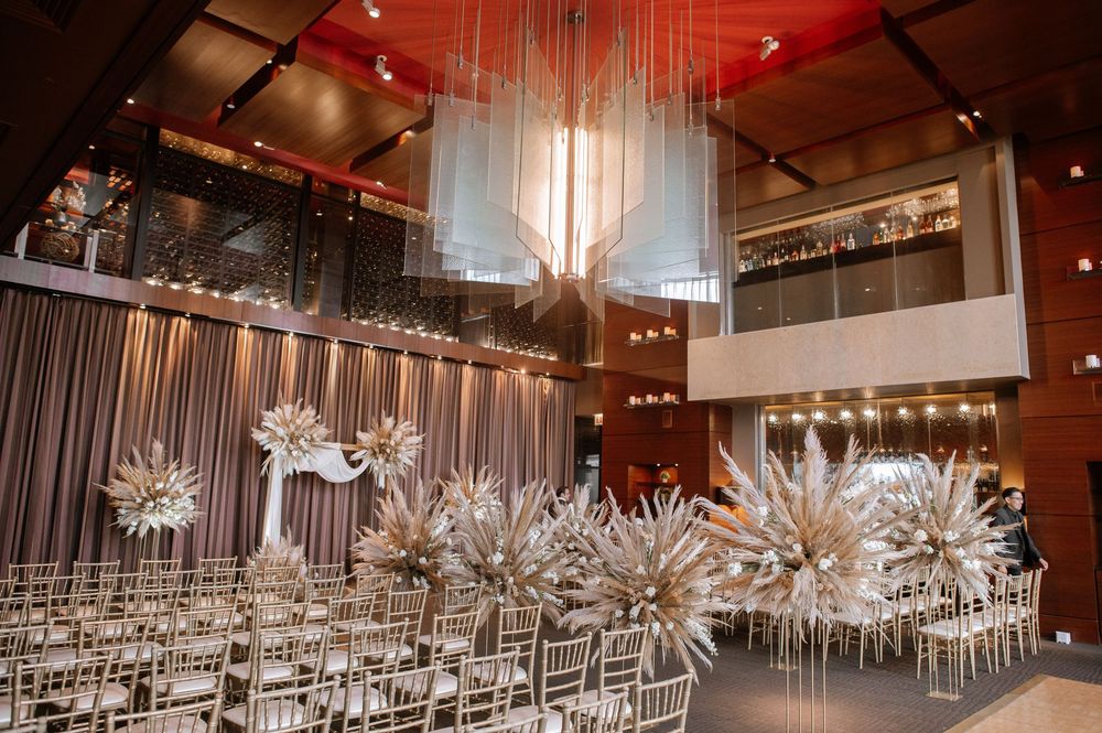 Modern wedding ceremony setup with dramatic lighting, silver ghost chairs, and ethereal pampas grass decorations in an industrial venue.