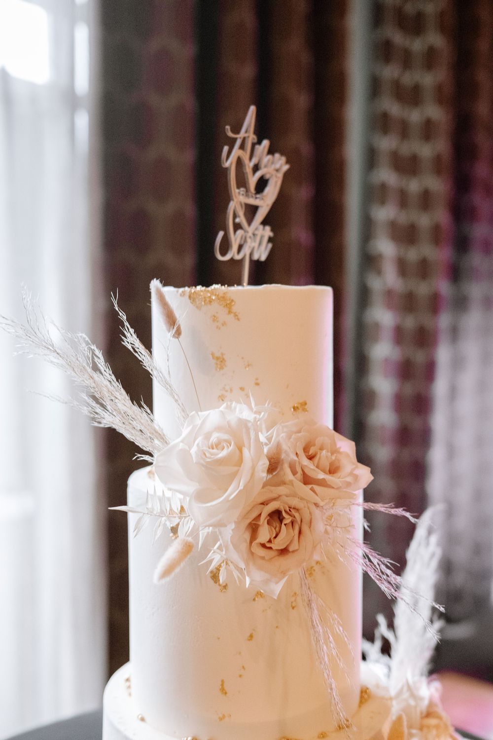 Elegant white wedding cake decorated with soft blush roses, wispy white feathers and a gold cake topper.