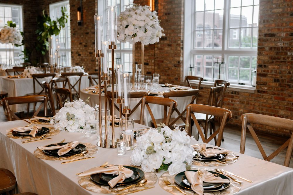 Elegant wedding reception table with tall white floral centerpieces, rustic wooden chairs, and exposed brick walls.