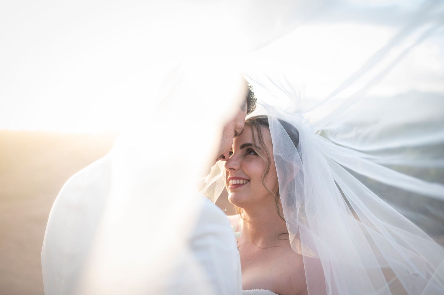 Beautiful bride in flowing white veil smiles radiantly during golden hour outdoor photo session.