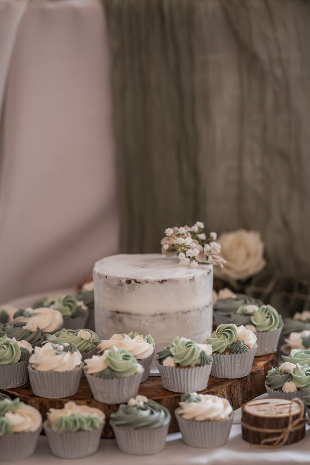 Elegant white wedding cake with sage green and cream frosted cupcakes displayed in a rustic setting.