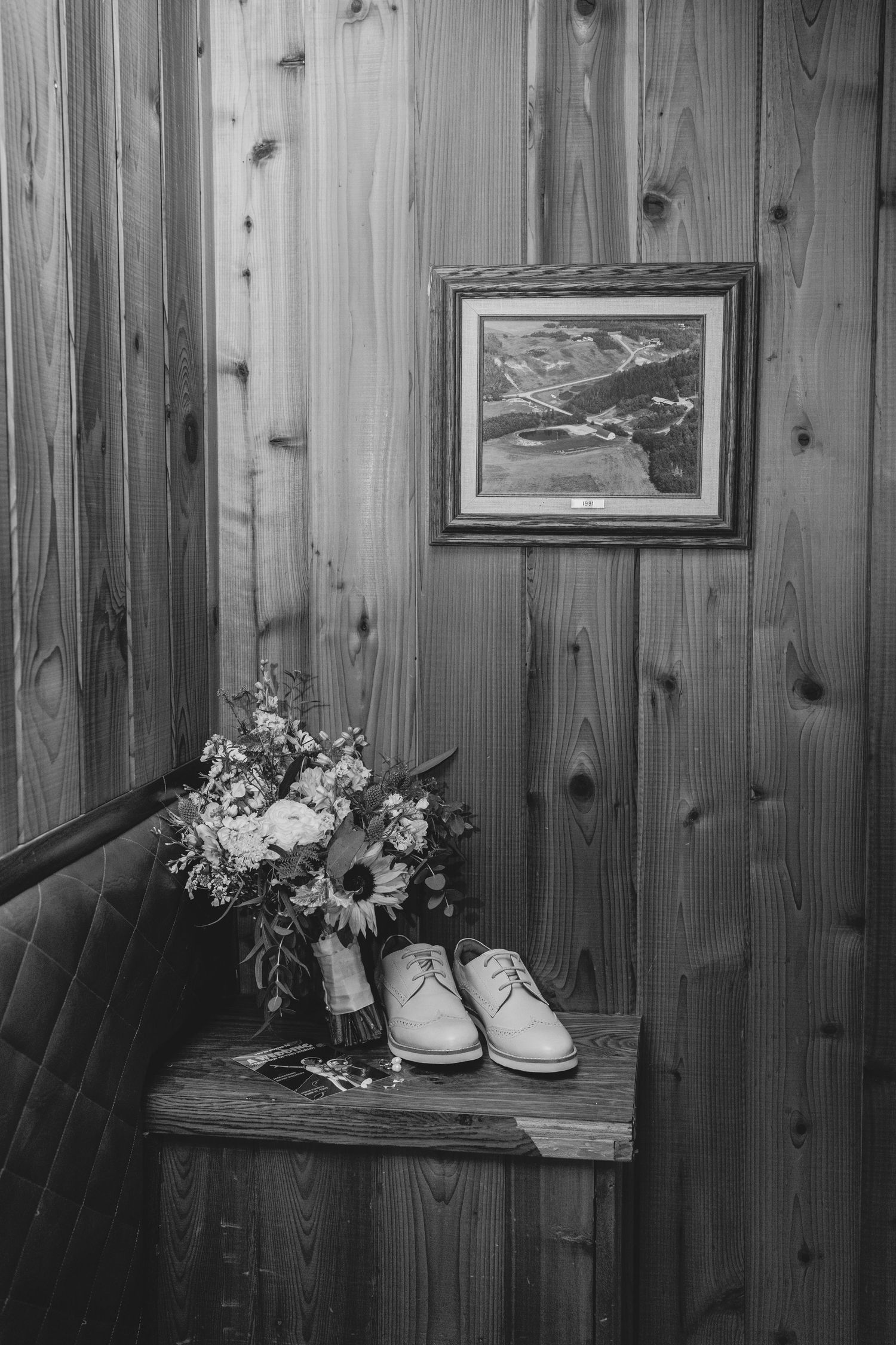 Black and white photo of rustic wooden wall decor with white shoes and floral bouquet on bench.