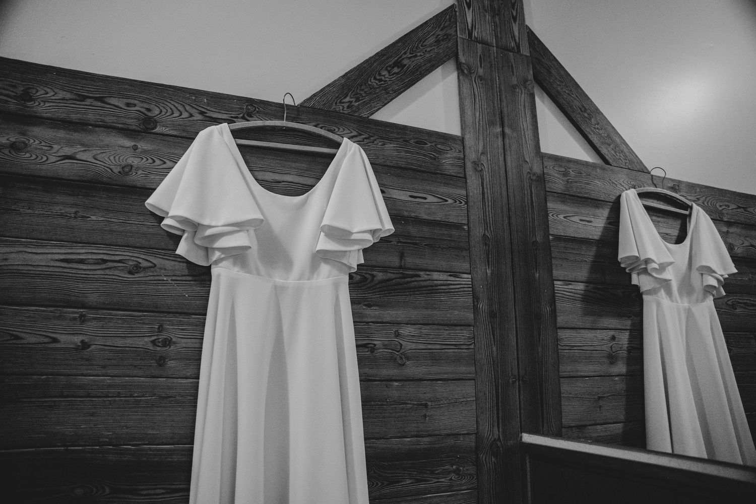 Two white wedding dresses hanging on wooden beams in black and white.