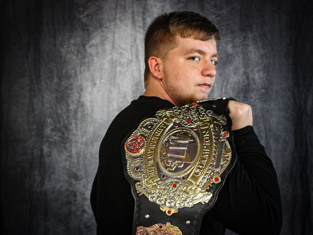 Professional wrestler displays championship belt against dark textured background.
