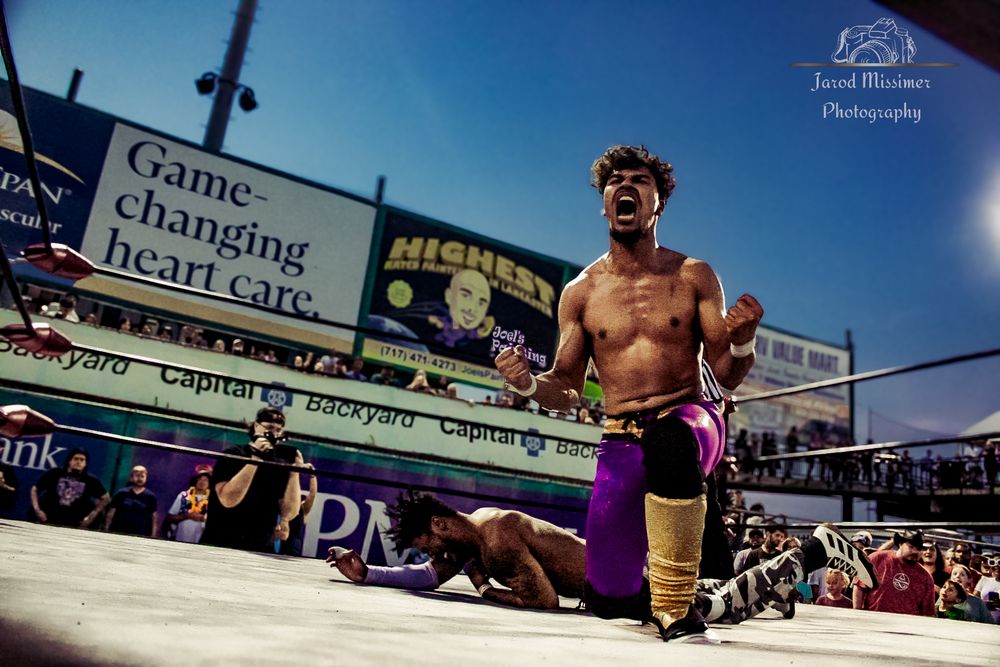 Professional wrestler performs in an outdoor ring with advertisement banner visible in background.