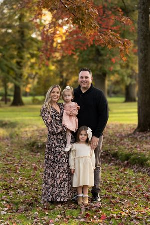 A family poses together on fallen autumn leaves under colorful fall foliage at sunset in a park.