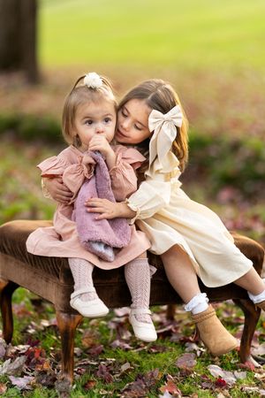 Two children sharing a tender moment while sitting on wooden benches surrounded by autumn leaves in a garden setting.